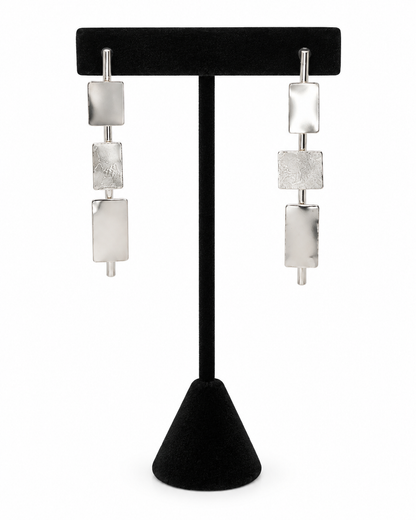 Clarity geometric sterling silver earrings featuring three rectangular metal elements arranged vertically on slim bars, displayed on a black jewelry stand.